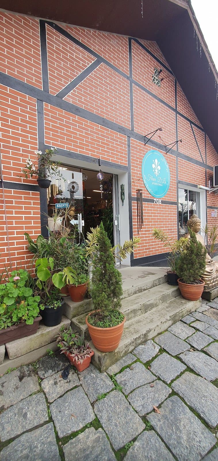 A fachada de enxaimel da Fleur D'Lis, de tijolo à vista e vigas escuras, com a placa redonda turquesa ao lado da porta e vasos de plantas na escada